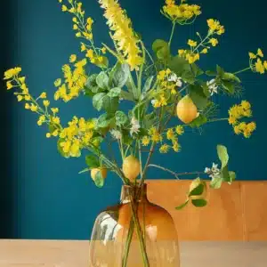 Jula Bloom’s Ghandi silk flower bouquet with yellow blooms and hanging lemons, in a semi opaque brown glass vase on a table