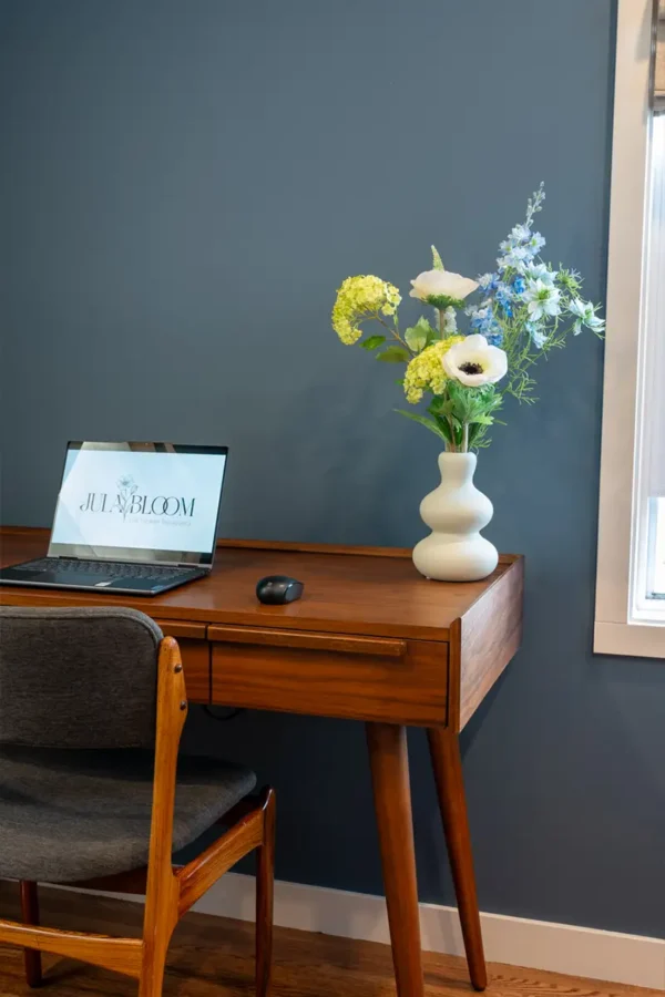 Jula Bloom’s Greta silk flower bouquet with white and blue blooms in a white vase, presented on a wooden desk.